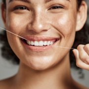Une jeune femme souriante utilise du fil dentaire, montrant des dents blanches et saines, illustrant l'importance de l'hygiène bucco-dentaire.