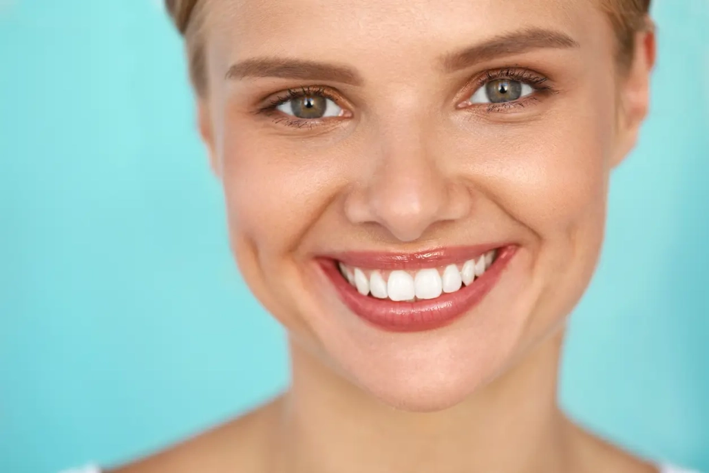 Portrait d'une femme souriante aux yeux verts et aux dents blanches parfaites, posant devant un fond bleu clair.